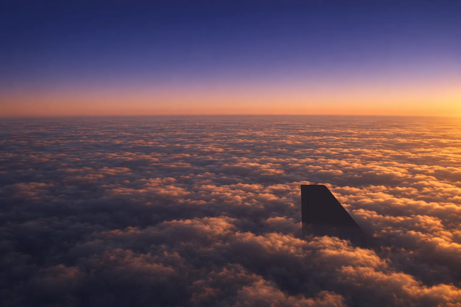 A deck of cloud seen from cruise altitude a few minutes after sunrise, lit amber along the tops under a deep indigo sky, the tail fin of an airliner cutting up through it