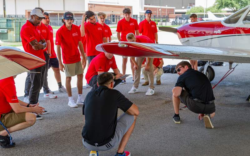 Thrust Flight students gathered around an instructor under a Piper Archer wing